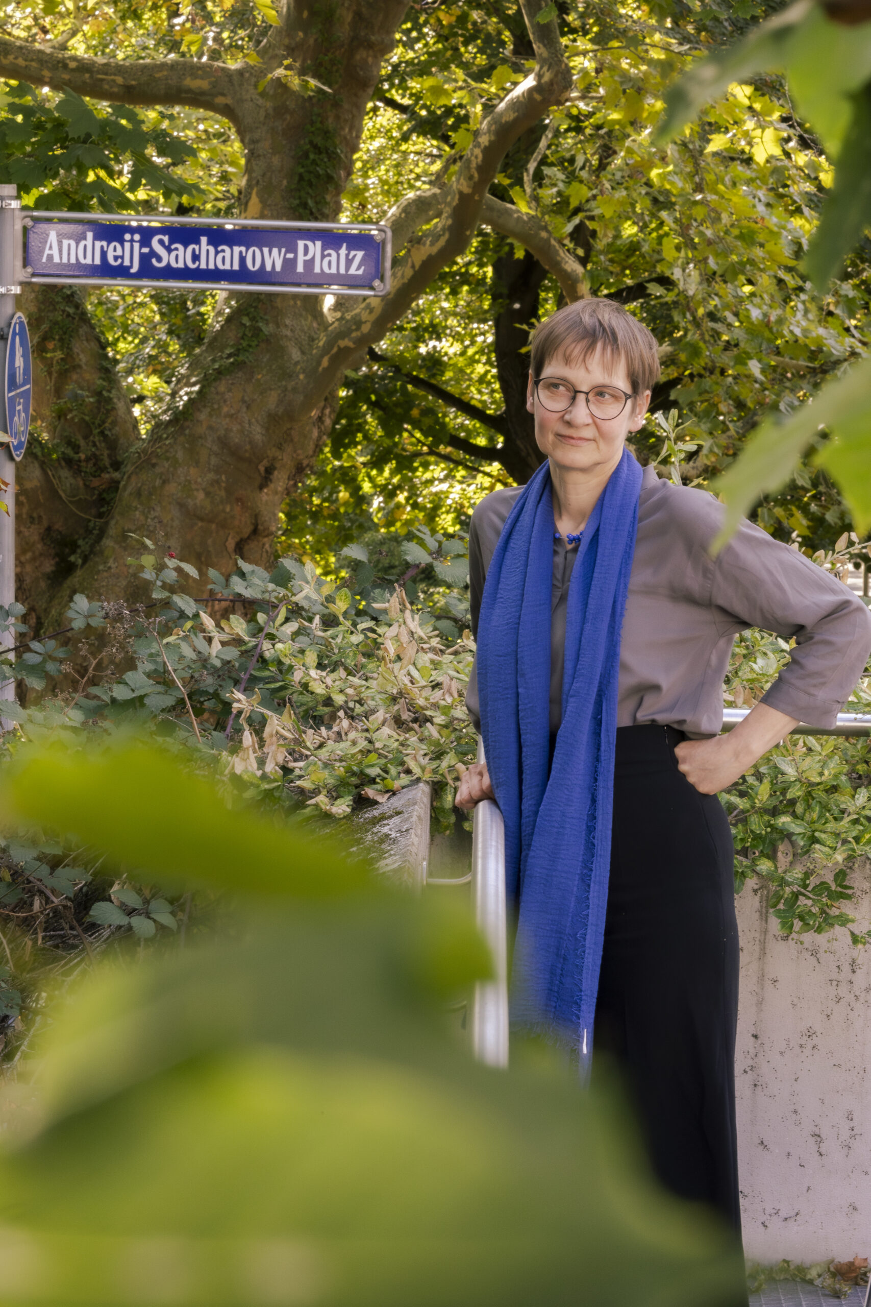 Prof. Dr. Eva Pils next to the Andreji-Sacharow-Platz sign surrounded by green.